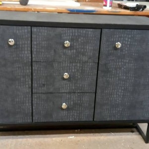 May include: A black sideboard with three doors and two drawers. The doors and drawers have a textured, crocodile-skin pattern. The sideboard has silver knobs and sits on a black metal frame.