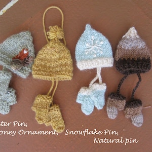 Può includere: Cinque berretti a maglia con guanti abbinati. I berretti sono di vari colori e motivi, tra cui marrone, bianco, blu e giallo. I guanti sono anche di vari colori e motivi. I berretti sono etichettati come "Forester Pin, Honey Ornament, Snowflake Pin, Natural Pin".