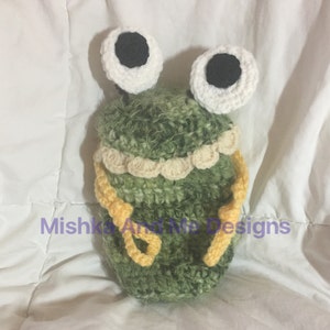 May include: A handmade crochet frog-shaped item in shades of green, white, and yellow. The frog has large white eyes with black pupils, a scalloped cream-colored trim, and yellow straps. The text "Mishka And Me Designs" is visible.