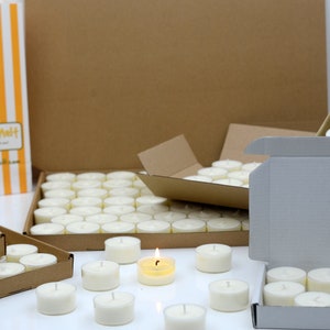 May include: A large quantity of white tea light candles are packaged in cardboard boxes. The boxes are open and the candles are visible. One candle is lit and burning.