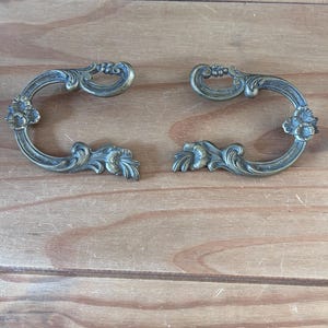 Peut inclure: Deux poignées de tiroir en métal couleur laiton assorties, présentant une forme en C ouverte et incurvée. Chaque poignée est ornée de motifs floraux en relief aux extrémités et le long du cadre incurvé, avec une finition métallique vieillie.