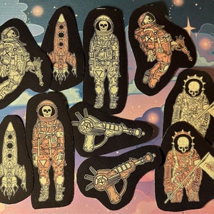 May include: Ten embroidered iron-on patches featuring a space exploration theme. The patches are black with white and pink outlines. The patches feature astronauts, spaceships, and ray guns.