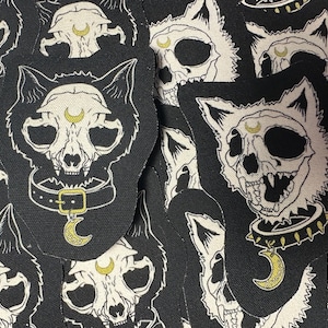 May include: Black and white patches featuring a cat skull design with a crescent moon and collar. The patches have a black background and a white outline, creating a striking contrast. The cat skull has a detailed design.