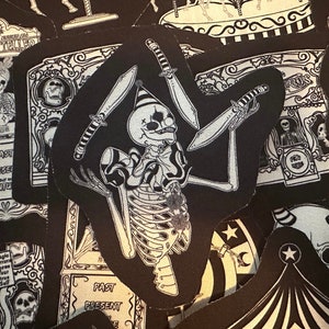 May include: A black and white illustration of a skeleton clown juggling knives. The clown is wearing a hat and a ruffled collar. The illustration is on a black fabric patch.