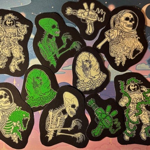 Op de afbeelding: Set van tien strijkbare patches met verschillende ruimtethema-illustraties. De patches zijn zwart met witte en groene details. De illustraties omvatten skeletten, aliens en astronauten.