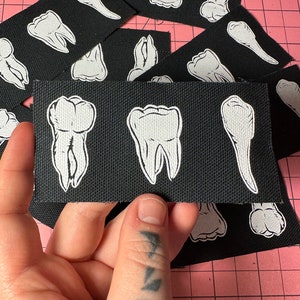 May include: A set of black fabric patches with white embroidered teeth. The patches are arranged in a grid pattern and feature different types of teeth, including molars, incisors, and canines.