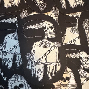 May include: A collection of black and white embroidered iron-on patches featuring various skeleton designs. The designs include a skeleton wearing a suit, a skeleton with a dripping head, and a skeleton in a pharaoh's headdress.