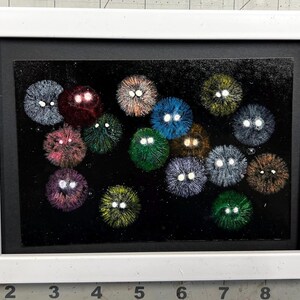 May include: A framed artwork featuring a black background with multiple colorful, fuzzy, orb-like shapes. Each orb has two white, eye-like details. The frame is white, and the artwork is mounted on a black mat.