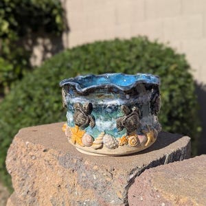 May include: A ceramic planter featuring a wavy rim and a glossy blue and white glaze. The exterior is decorated with three-dimensional sculpted sea turtles and various seashells in shades of tan, orange, and white along the base.