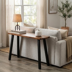May include: A narrow console table with a smooth wooden top and four black metal legs. A table lamp with a textured beige shade and a white ceramic vase are placed on the surface. The table is positioned behind a light gray upholstered sofa.