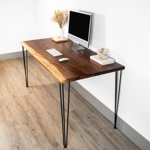 May include: A dark brown wooden desk with a live edge and black hairpin legs. A computer monitor, keyboard, and mouse sit on the desk. A vase with dried plants and decorative items are also on the desk.