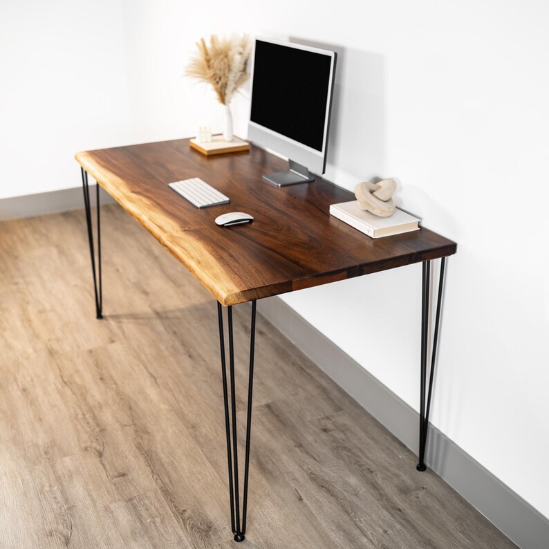 May include: A dark brown wooden desk with a live edge and black hairpin legs. A computer monitor, keyboard, and mouse sit on the desk. A vase with dried plants and decorative items are also on the desk.
