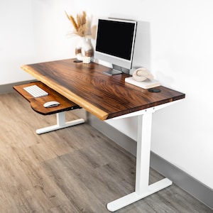 May include: A white standing desk with pull-out keyboard tray. The adjustable height desk has a white base. The wooden top is a rich brown color with a natural edge. The keyboard tray is made of wood and has a white keyboard and mouse on it.