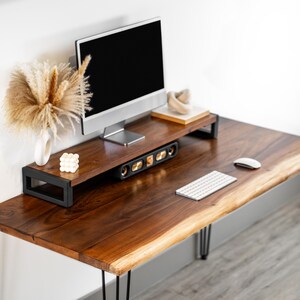 May include: A walnut wood desk with a live edge top and black hairpin legs. A computer monitor is on a black metal stand with a speaker system underneath. A white wireless keyboard and mouse are on the desk.
