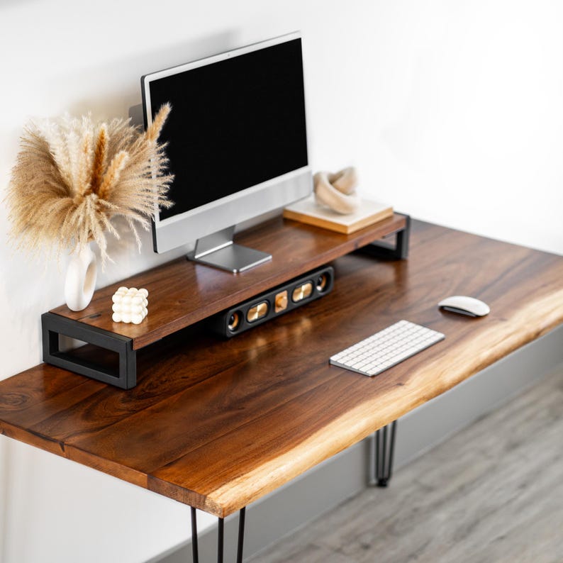 May include: A walnut wood desk with a live edge top and black hairpin legs. A computer monitor is on a black metal stand with a speaker system underneath. A white wireless keyboard and mouse are on the desk.