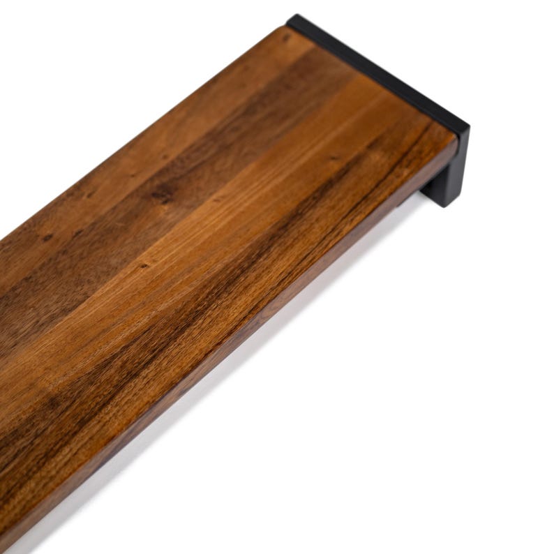 May include: A close-up of a wooden desk shelf with a dark brown stain. The shelf has a black metal bracket on the right side.