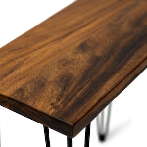 May include: Close-up of a walnut end table with a dark brown wood top and black hairpin legs. The living room table is made of solid wood and has a natural finish.