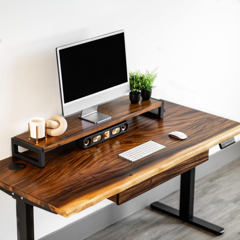 May include: A wooden standing desk with a black metal frame and a drawer. The live-edge desk has a walnut wood top with a natural edge and a monitor stand with a speaker system and a computer monitor on top. A keyboard and mouse are on the desk.