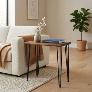 May include: A walnut side table with black hairpin legs. The small end table has a brown wood top and is decorated with a book and a vase. The small living room table is next to a white couch.