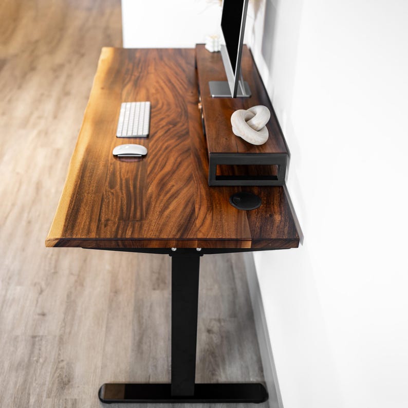 May include: A wood standing desk with a black metal sit-stand frame and a large, dark wood desktop. The desktop has a natural wood grain pattern and is accented with a black metal monitor shelf. A white keyboard and mouse are on the desktop.