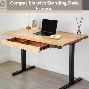 May include: A light-colored wooden standing desk with a black frame. The desk has a drawer that is partially open, revealing two compartments. A laptop, notepad, and lamp are on the desk. The text "Compatible with Standing Desk Frames" is at the top.