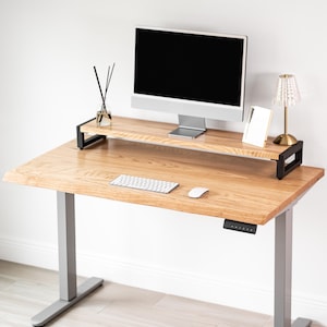 May include: A white oak wood standing desk with a black monitor stand, a silver computer monitor, and a white keyboard and mouse. A small lamp and a diffuser are on the desk. The desk has gray legs.