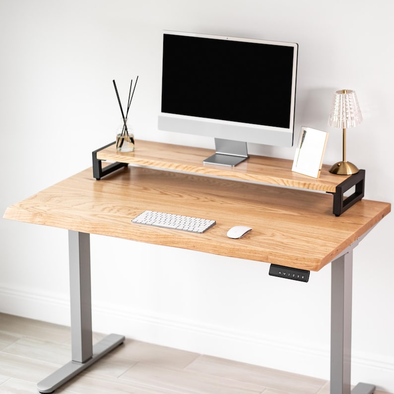 May include: A white oak wood standing desk with a black monitor stand, a silver computer monitor, and a white keyboard and mouse. A small lamp and a diffuser are on the desk. The desk has gray legs.