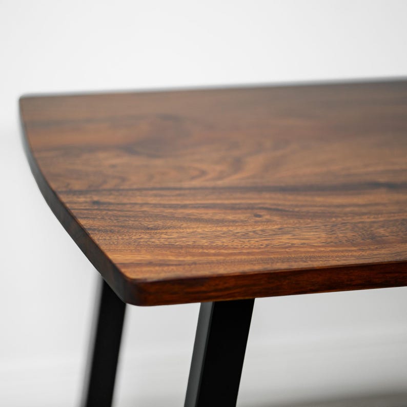 May include: A close-up of a wooden dining table with a dark walnut surface and black legs. The kitchen table has a curved edge and a natural wood grain pattern. The legs are angled and provide support. The table is likely used for home decor or furniture.