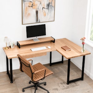 May include: An L-shaped wooden desk with a monitor, keyboard, and mouse. The desk has black metal legs and a brown leather office chair. A monitor stand, a small lamp, and a framed picture are on the desk. A notebook and a vase of flowers are also visible.