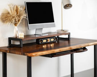 May include: A dark wood desk with black metal legs, a monitor, and a speaker. A monitor stand and a keyboard tray are attached to the desk. A white lamp and decorative vase with dried flowers are on the desk.
