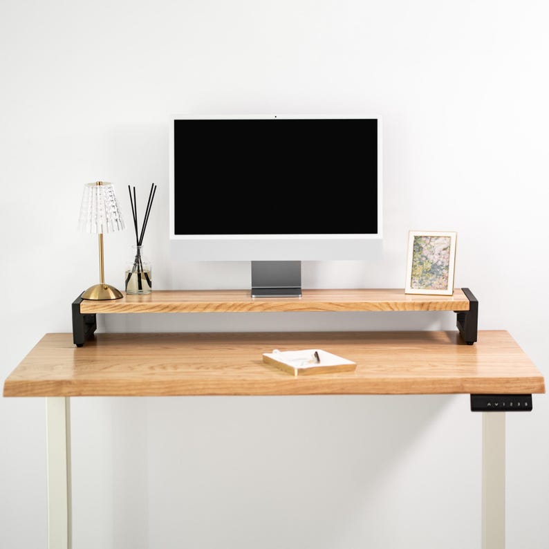 May include: A wooden standing desk with a monitor, lamp, and picture frame. The desk has a light wood finish and black metal supports. A small tray and diffuser are on the desk. The monitor is black.