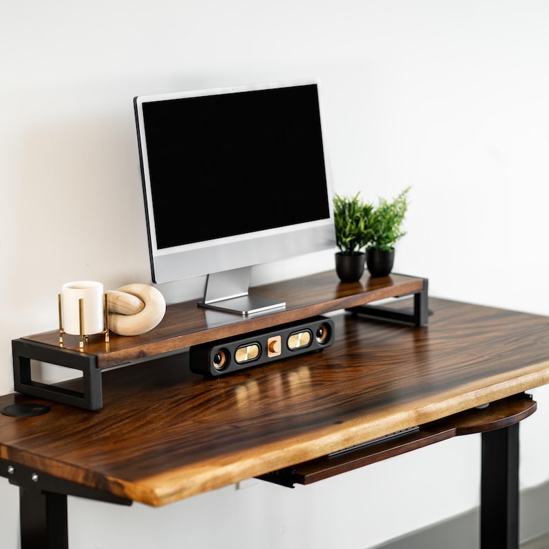 May include: A wooden standing desk with a black metal adjustable frame and a monitor stand. The wooden desk has a live edge and a built-in drawer. A computer monitor, a speaker system, and decorative items are on the desk.