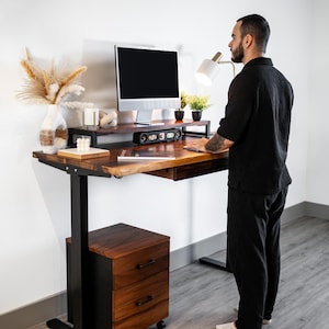 May include: A standing desk with live edge and black height adjustable legs. The desk has a built-in drawer and a matching rolling file cabinet. A computer monitor, speakers, and decorative items sit on the desk. A person is standing at the desk.