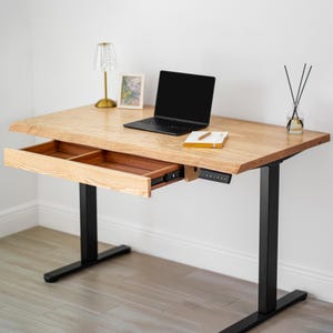 May include: A white oak standing desk with a drawer open. The desk has a black laptop, a small lamp, a picture frame, and a notebook on top. The desk is supported by black metal legs.