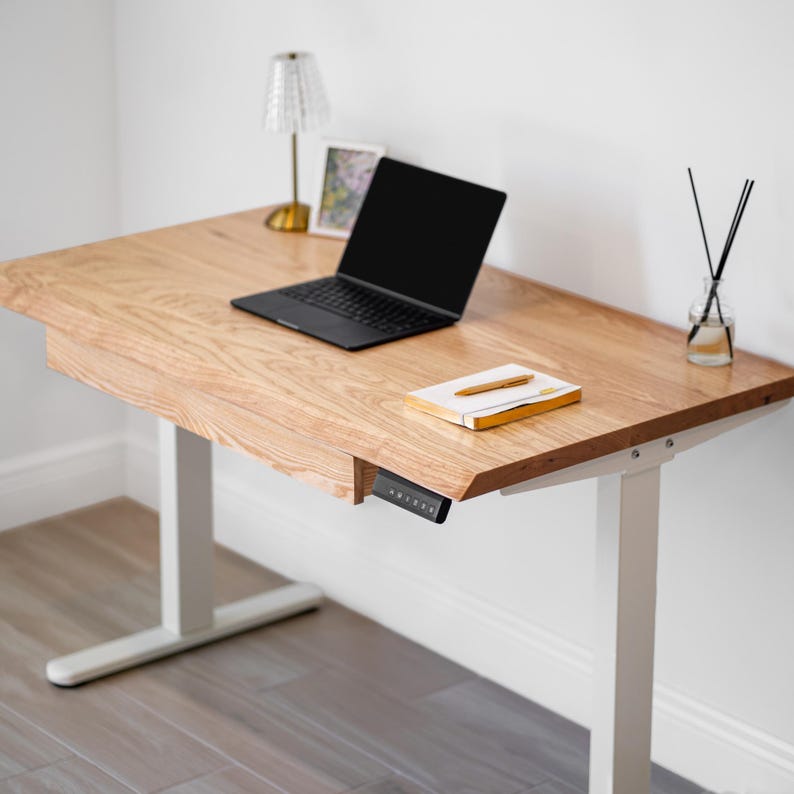 May include: A white oak desk with drawer, a black laptop, a notebook, and a pen. The desk has white legs and a control panel. A small lamp, a framed picture, and a diffuser are also on the desk.