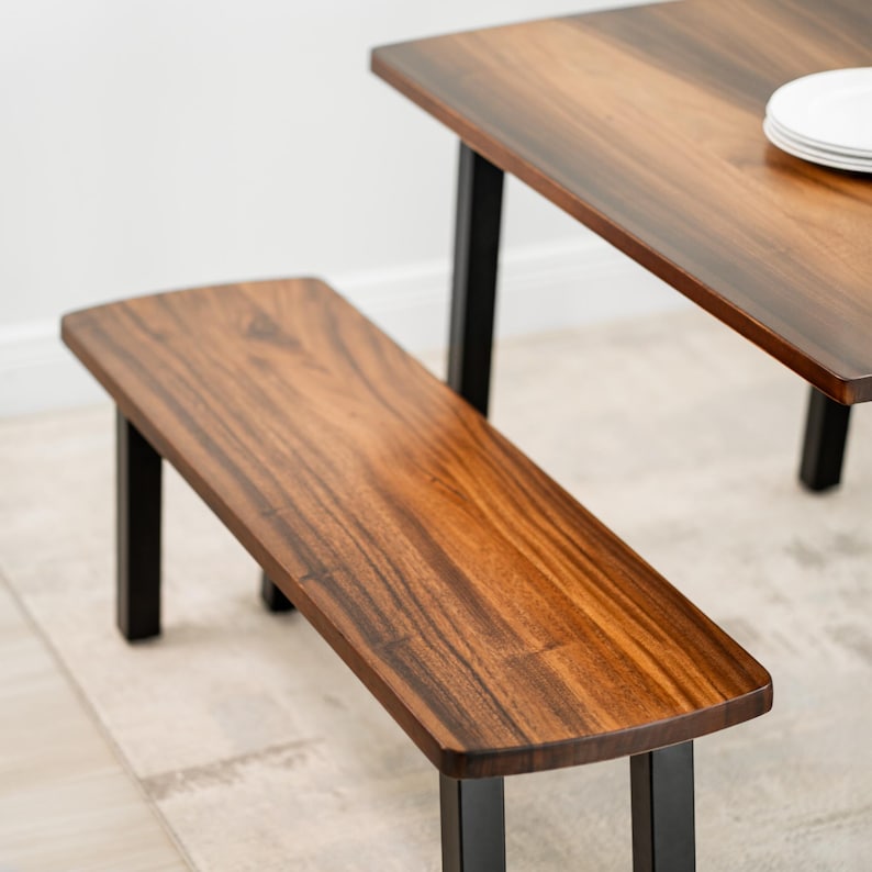May include: A solid-wood dining table with a wooden bench in a dark brown finish and rounded edges. The bench is supported by black metal legs. A matching table with a similar finish and black legs is partially visible in the background. The table has white plates on top.