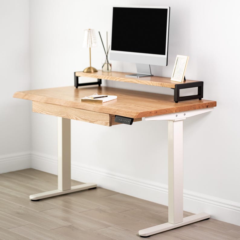 May include: A white oak adjustable standing desk with a monitor shelf and a drawer. The desk has white legs and a black control panel. A small lamp and a picture frame are on the desk.