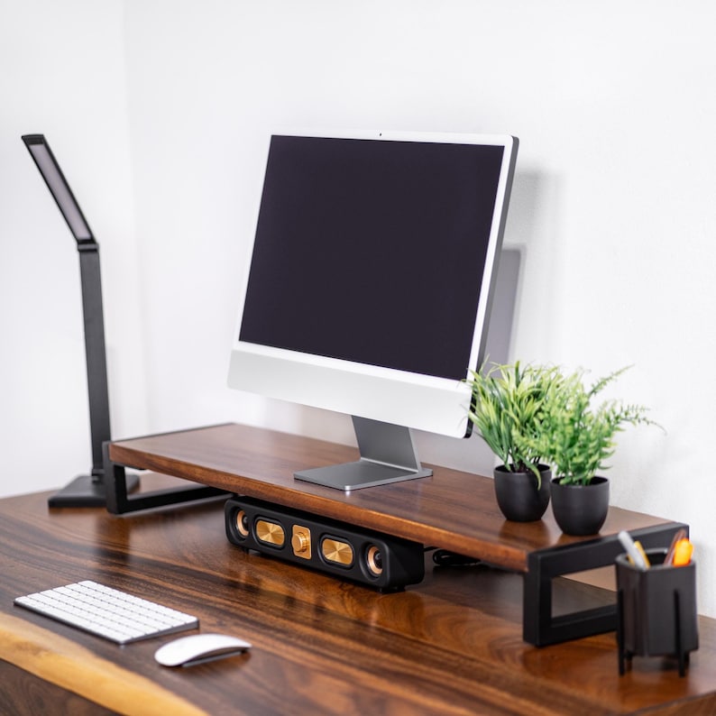May include: A wooden computer monitor stand with a black metal frame. The riser monitor stand has a speaker system with gold accents. A computer monitor, two potted plants, and a desk lamp are on the stand.