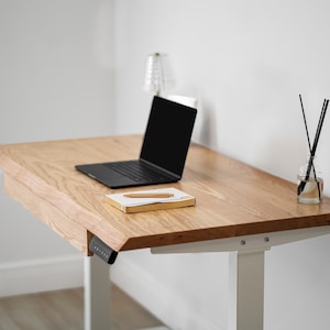 May include: A white oak standing desk with drawer, a black laptop, a gold pen, and a notebook. A glass diffuser with black reeds and a small lamp are also on the desk. The desk has white legs and a black control panel.