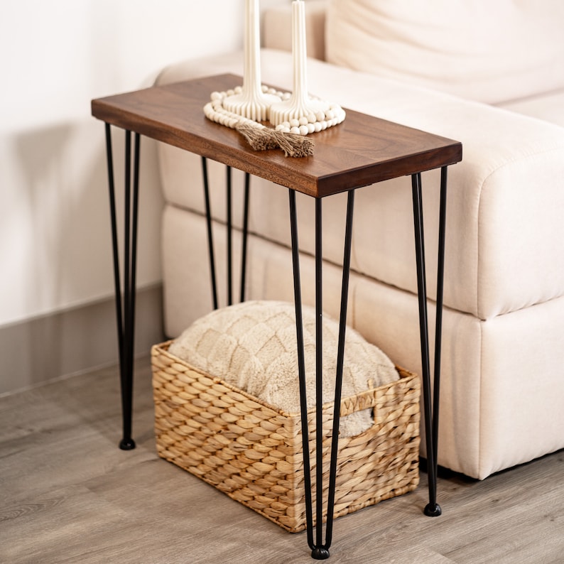 May include: A walnut side table with black hairpin legs. The small end table has a woven basket underneath and a white beaded garland on top.