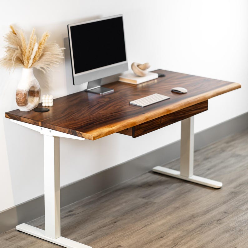 May include: A walnut standing desk with a wooden top and a single drawer. The standing desk has a white metal frame and a dark wood top with a live edge. A computer monitor, keyboard, and mouse are on the desk.