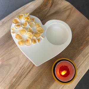 Op de afbeelding: Een witte hartvormige kaarsenhouder met een groep driedimensionale bloemen met goudkleurige centra aan de ene kant en een ronde uitsparing voor een waxinelichtje aan de andere kant, vergezeld door een kleine gele houder met een brandend rood kaarsje.