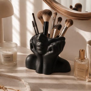 Peut inclure: Un porte-pinceaux noir en forme de visage rempli de divers pinceaux. Le support est posé sur une surface en marbre, à côté d'une bougie, d'un flacon de parfum et d'un plateau décoratif avec des bijoux. Un miroir rond est en arrière-plan.