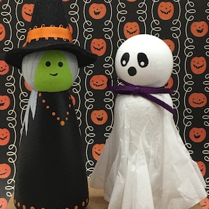 May include: A black and green witch doll with an orange hat and a white ghost doll with a purple bow. Both dolls are standing on a brown surface with a background of orange pumpkins on a black and white pattern.