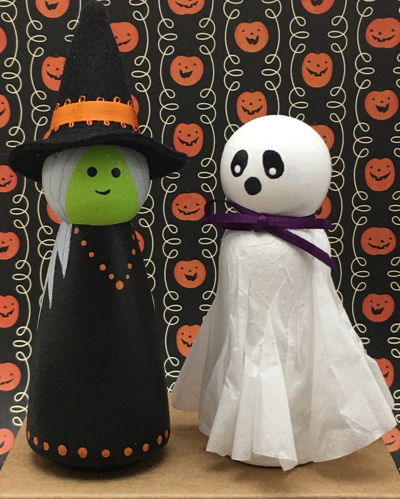 May include: A black and green witch doll with an orange hat and a white ghost doll with a purple bow. Both dolls are standing on a brown surface with a background of orange pumpkins on a black and white pattern.