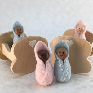 May include: Three wooden cribs with felt baby dolls inside. The cribs are painted a light wood color and have white hearts painted on the side. The dolls are wearing blue and pink polka dot outfits.