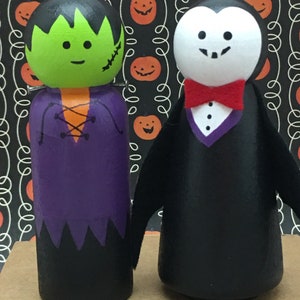 May include: Two wooden peg dolls dressed as Halloween characters. The doll on the left is Frankenstein's monster, with green skin, black hair, and a purple shirt. The doll on the right is a vampire, with a black suit, white face, and a red bow tie.