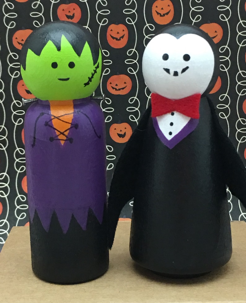 May include: Two wooden peg dolls dressed as Halloween characters. The doll on the left is Frankenstein's monster, with green skin, black hair, and a purple shirt. The doll on the right is a vampire, with a black suit, white face, and a red bow tie.