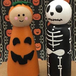 May include: Two wooden peg dolls dressed up for Halloween. One doll is painted to look like a pumpkin with a green stem and a friendly face. The other doll is painted to look like a skeleton with a black body and white bones.