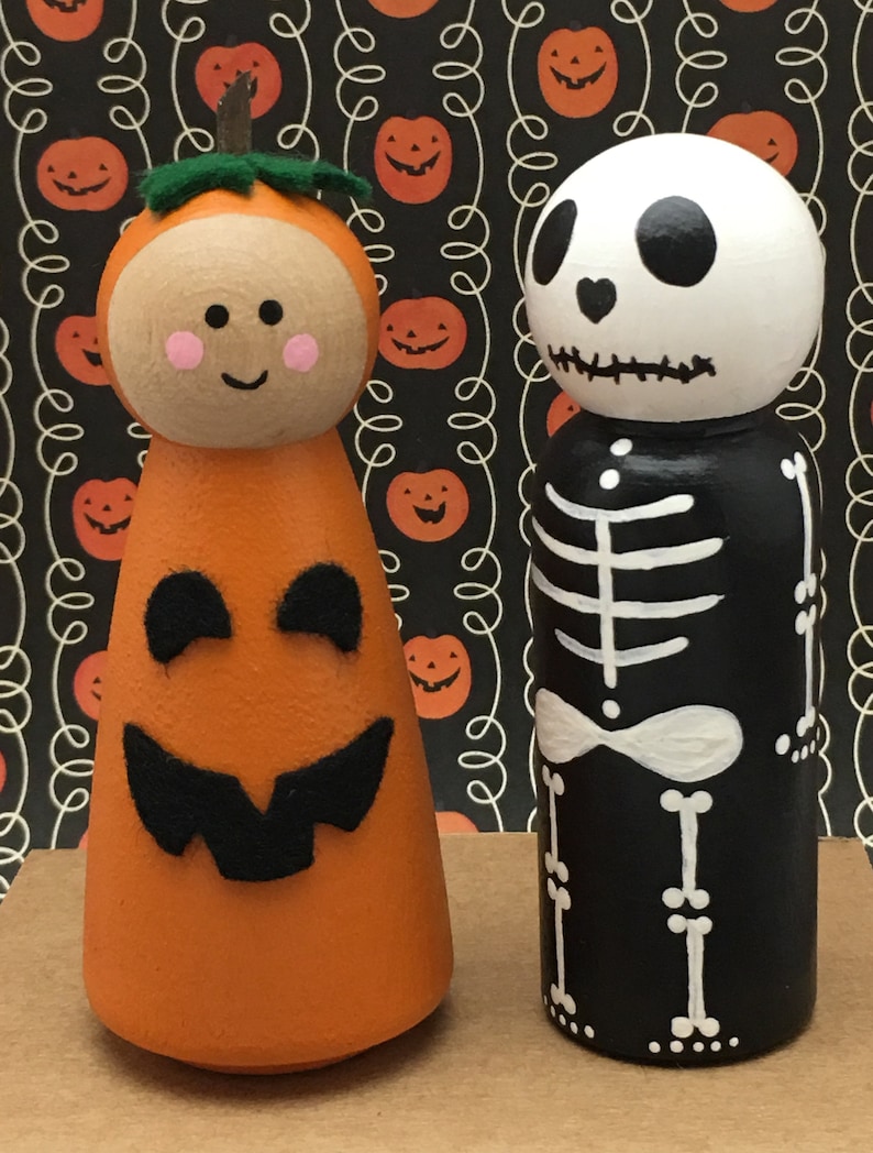 May include: Two wooden peg dolls dressed up for Halloween. One doll is painted to look like a pumpkin with a green stem and a friendly face. The other doll is painted to look like a skeleton with a black body and white bones.